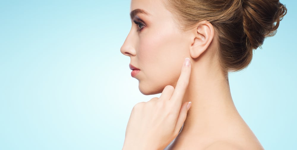 A beautiful young woman points to her ear against a blue background, highlighting the area considered for cosmetic earlobe repair treatment.