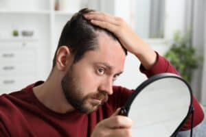 Man with hair loss problem looking in mirror indoors, considering FUE and FUT hair transplant treatments for hair restoration